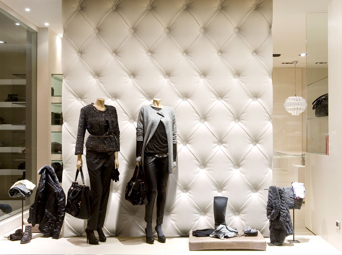 Escaparate de tienda de moda con maniquíes vistiendo ropa elegante con mucho color, junto a accesorios de moda sobre un fondo de pared acolchada clara.
