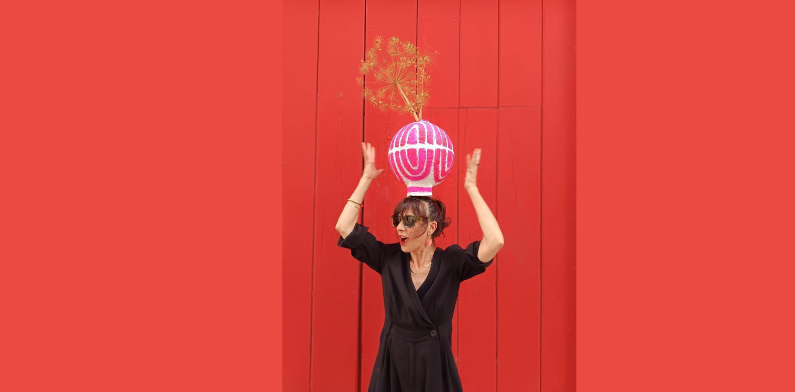 Una mujer con un vestido negro con mucho  Diseño, sostiene un objeto colorido en su cabeza mientras está frente a un fondo rojo.